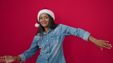 Genç Afrikalı Amerikalı kadın Noel şapkası takıp kırmızı arka planda dans ediyor.