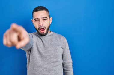 Mavi arka planda dikilen İspanyol adam parmağı önde şaşırmış, ağzı açık şaşkınlık ifadesi, ön tarafta bir şey var. 