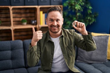 Orta yaşlı beyaz bir adam elinde sanal para birimiyle gülümsüyor mutlu ve pozitif gülümsüyor, başparmağıyla mükemmel ve onay işareti yapıyor. 