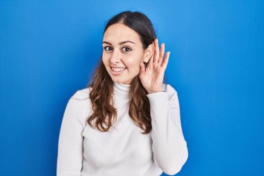 Young hispanic woman standing over blue background smiling with hand over ear listening an hearing to rumor or gossip. deafness concept. 
