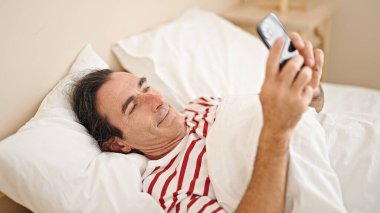 Orta yaşlı bir adam yatakta uzanmış, yatakta gülümsüyor.