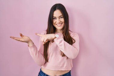 Young brunette woman standing over pink background amazed and smiling to the camera while presenting with hand and pointing with finger. 