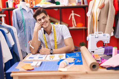 Young hispanic man tailor smiling confident sitting on table at atelier
