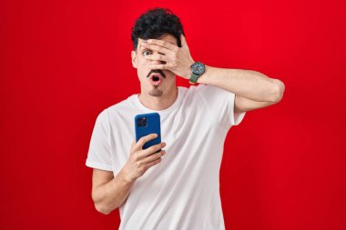 İspanyol adam kırmızı arka planda akıllı telefon kullanıyor. Şoka girip yüzünü ve gözlerini kapatıyor. Utanmış bir ifadeyle parmaklarından bakıyor.. 