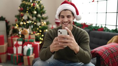 Young hispanic man using smartphone sitting on sofa by christmas tree at home