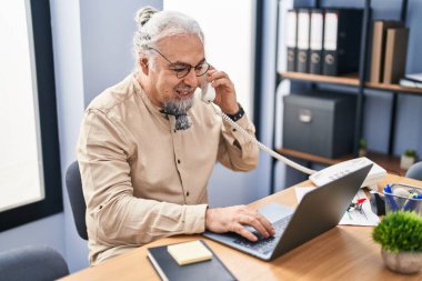 Middle age grey-haired man business worker talking on telephone using laptop at office