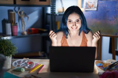 Mavi saçlı genç modern kız, geceleri dizüstü bilgisayarla sanat stüdyosunda oturuyor. Kollarını kaldırıp, gözlerini açarak başarıyı kutluyor ve şaşırıyor. Kazanan konsept. 