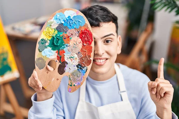 Non binary person holding palette close to face smiling with an idea or question pointing finger with happy face, number one