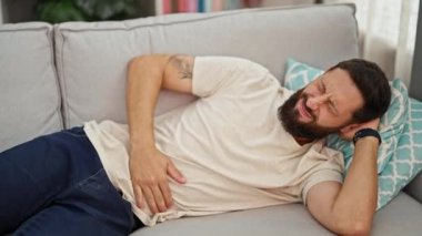 Başı ağrıyan genç adam kanepede uzanıyor ve evde televizyon seyrediyor.