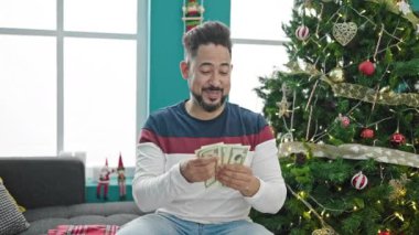 Yakışıklı, sakallı, süveterli genç adam Noel zamanı oturma odasında para sayıyor.