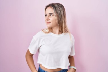 Pembe arka planda duran genç sarışın kadın yüzünde gülümseme ve doğal ifadeyle yan yana bakıyor. Kendinden emin gülen. 