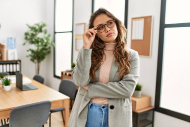 Genç, güzel, İspanyol bir iş kadını. Kollarını kavuşturup ofiste duruyor.