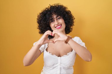 Sarı arka planda duran kıvırcık saçlı, genç esmer kadın elleriyle kalp sembolü yapan aşka gülümsüyor. romantik kavram. 