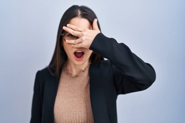 Genç esmer kadın mavi arka planda dikiliyor. Şoka girip yüzünü ve gözlerini kapatıyor. Utanmış bir ifadeyle parmaklarından bakıyor.. 