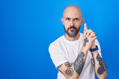 Mavi arka planda dikilen İspanyol dövmeli bir adam. Elinde sembolik bir silahla el hareketleriyle cinayet silahlarıyla oynuyor. Kızgın bir yüz. 