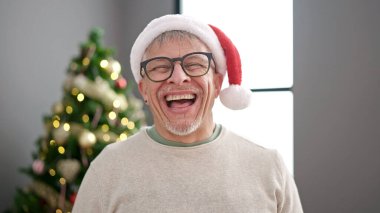 Orta yaşlı, kır saçlı, gülümseyen, kendine güvenen bir adam Noel ağacının yanında duruyor.