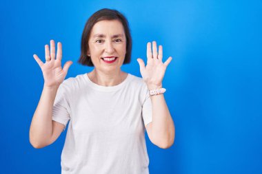 Orta yaşlı İspanyol kadın mavi arka planda duruyor ve parmaklarını on numarayı gösterip işaret ederken kendinden emin ve mutlu gülümsüyor.. 