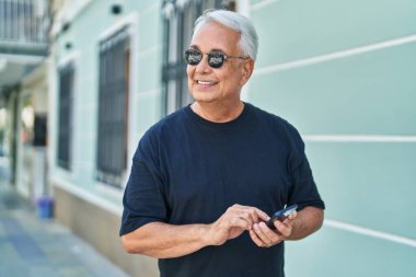 Orta yaşlı, kır saçlı, kendine güvenen, caddede akıllı telefon kullanan bir adam.