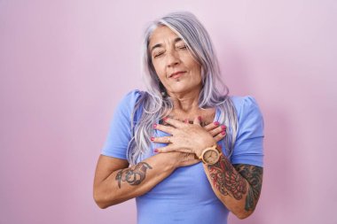 Pembe arka planda duran, elleri göğsünde, gözleri kapalı ve yüzünde minnet ifadesi olan orta yaşlı bir kadın. sağlık kavramı. 