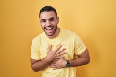 Sarı arka planda dikilen Latin asıllı genç adam gülümseyerek kahkahalarla gülüyor.. 