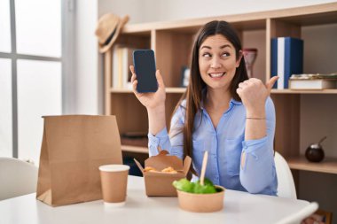 Genç esmer kadın evde yemek yiyor. Akıllı telefon ekranı gösteriyor. Başparmağıyla yan tarafı gösteriyor. 