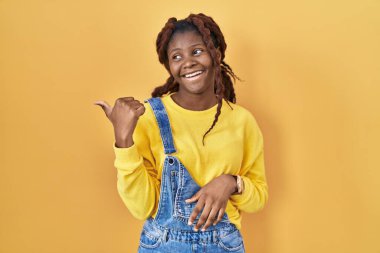 Sarı arka planda dikilen Afrikalı kadın gülümsüyor, gülümsüyor ve başparmağıyla yan tarafa bakıyor.. 