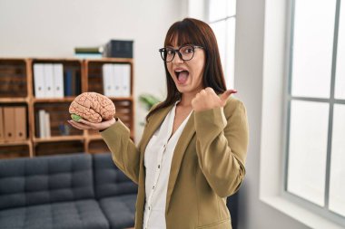 Terapi ofisinde çalışan genç bir terapist beyni tutup baş parmağını yana kaldırıyor ve ağzı açık gülümsüyor. 