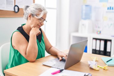 Ofiste çalışırken dizüstü bilgisayar kullanan, orta yaşlı, kır saçlı iş kadını.