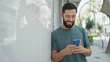 Sokak ortasında gülümseyen akıllı telefon ve kulaklık kullanan genç bir İspanyol.