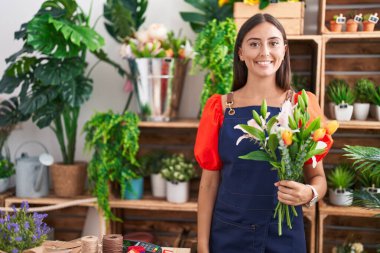 Young beautiful hispanic woman florist holding bouquet of flowers at florist