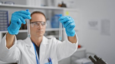 Hispanik orta yaşlı bir adam, ciddi bir bilim adamı, deneyine dalmış, laboratuarında test tüplerini güvenli bir şekilde tutuyor, tıbbi araştırmalar için sıvı örneklerini yoğun bir konsantrasyonla dikkatlice ölçüyor..