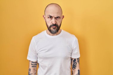 Sarı arka planda duran İspanyol dövmeli genç adam şüpheci ve gergin, sorun yüzünden surat asıyor. negatif kişi. 