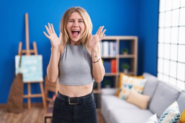 Sarışın, beyaz bir kadın oturma odasında durup zaferi kutluyor mutlu bir gülümseme ve ellerini kaldırarak kazanan bir ifadeyle. 