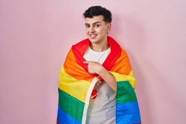 İkili olmayan kişi elinde gökkuşağı lgbtq bayrağı tutarken mutlu bir şekilde el ve parmağıyla işaret ediyor. 