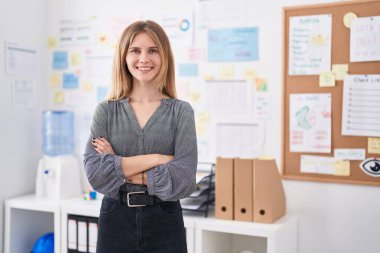 Genç, sarışın, iş kadını, gülümseyen, kendine güvenen, kollarını kavuşturup ofiste el kol kola duran.