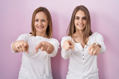 Orta yaşlı anne ve küçük kızı pembe arka planda dikiliyorlar. Seni ve kamerayı parmaklarıyla gösteriyorlar. Mutlu ve mutlu gülümsüyorlar. 