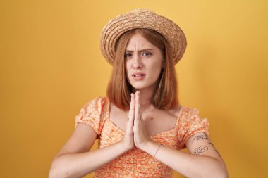 Sarı arka planda duran genç kızıl saçlı kadın yaz şapkası takıyor ve elleri ile yalvarıyor ve dua ediyor. Yüzünde umut ifadesi var. Çok duygusal ve endişeli. Yalvarıyor. 