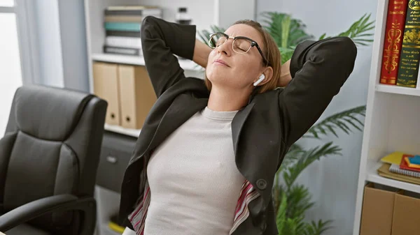 Relaxed young blonde business woman blissfully tuning into her favorite music at the office, hands cradling her head, immersing in the sound, forgetting the hustle of work life awhile.