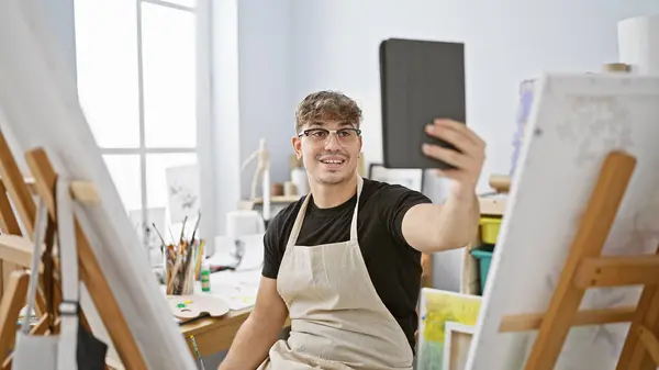 Charming young hispanic man, a budding artist, radiating joy while having a lively video call on his touchpad amidst the creative chaos of his art studio.