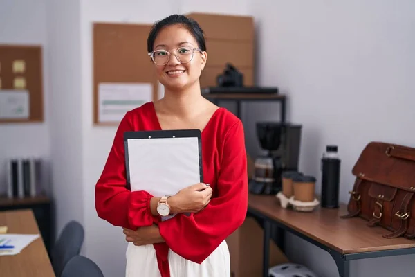 Young chinese woman business worker smiling confident holding clipboard at office