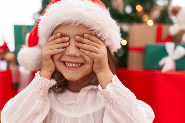 Adorable hispanic girl covering eyes with hands sitting on floor by christmas tree at home