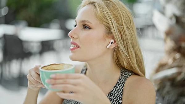 Young blonde woman drinking cup of coffee listening to music at coffee shop terrace