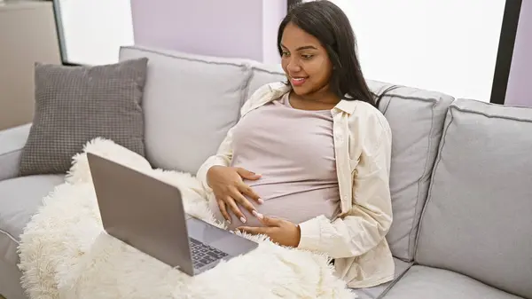 年轻貌美的孕妇 享受着母性的休息 舒适地在家里休息 按摩着肚子 在笔记本电脑屏幕上积极地微笑着 — 图库照片