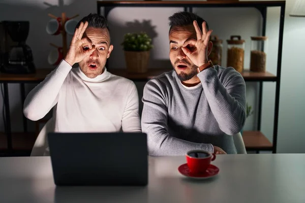 Homosexual couple using computer laptop doing ok gesture shocked with surprised face, eye looking through fingers. unbelieving expression.