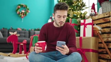 Genç adam Noel 'i evde akıllı telefon kullanarak ve çay içerek kutluyor.