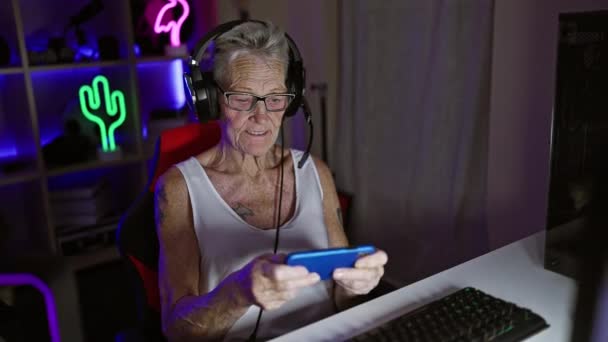 漂亮的白发苍苍的老年女性流言者在家里的游戏室里 在智能手机上玩电子游戏 沉醉于虚拟娱乐 中年但对数字了如指掌 — 图库视频影像