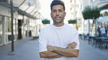 Gülümseyen İspanyol genç adam, güneşli şehir sokaklarında neşeli bir ifade olan çapraz kollarla durarak güven yayıyor.