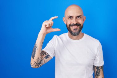 Mavi arka planda dikilen İspanyol dövmeli adam gülümseyen ve el hareketleriyle hareket eden parmakları küçük gösteren ve kameraya bakan adam. ölçüm kavramı. 