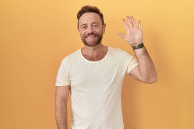 Sarı arka planda sakallı orta yaşlı bir adam beş numaralı parmaklarıyla işaret ederken kendinden emin ve mutlu bir şekilde gülümsüyor.. 