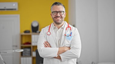 Gri saçlı doktor gülümsüyor. Kollarını kavuşturup klinikte duruyormuş.